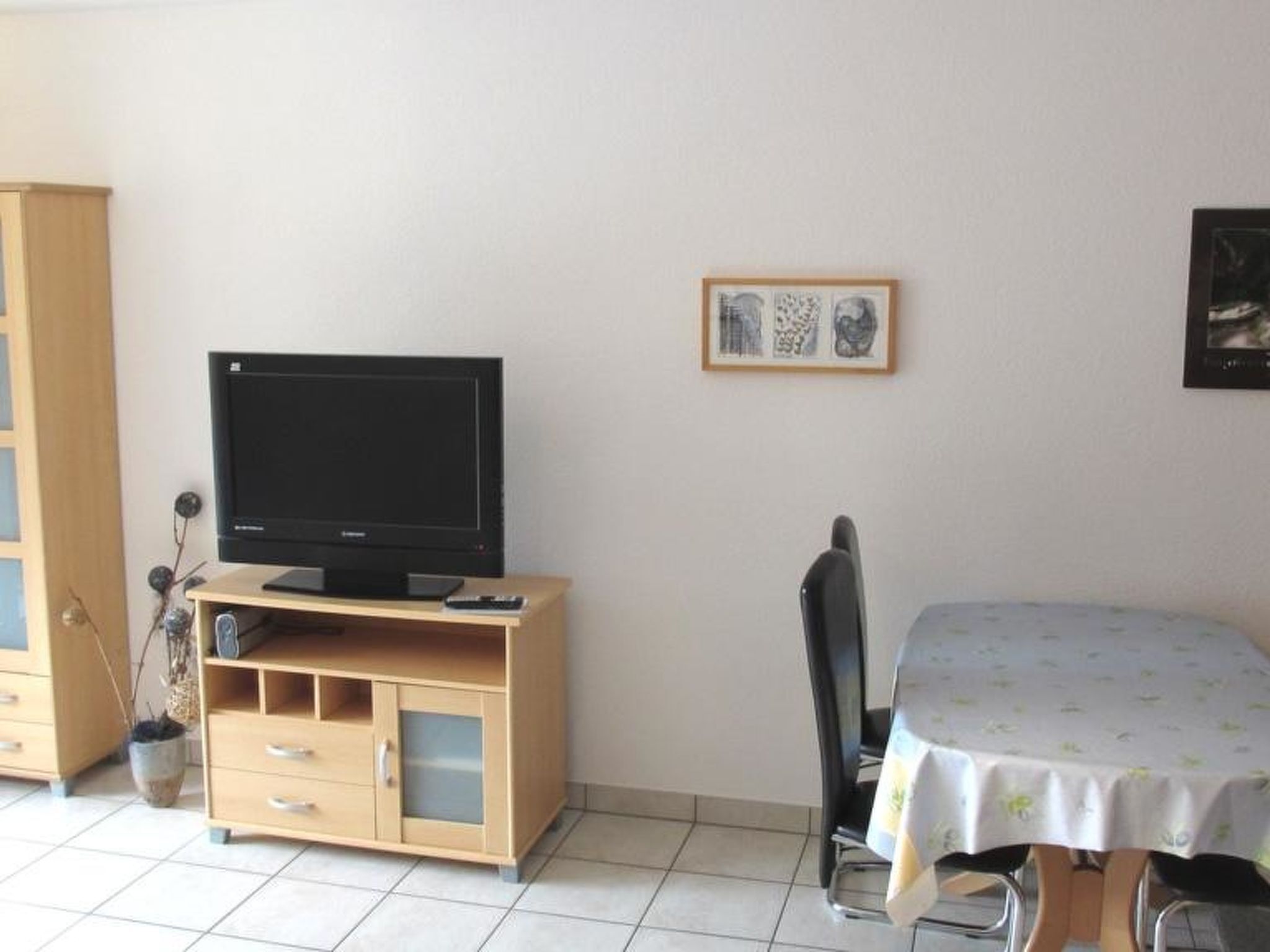 A room with a television on a wooden stand, a dining table with a tablecloth, a chair, and a cabinet with drawers.