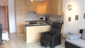 Un espacio habitable compacto con kitchenette, una silla de cuero negro, un sofá gris y una pequeña mesa de comedor.