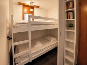 A bunk bed with a white headboard, a small bedside table, and a shelf with books and a potted plant. - Residenz Christina Leukerbad # 5-bed apartment, Dusche, WC by Interhome (Leukerbad)