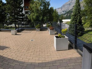 A paved courtyard with rectangular planters, a metal fence, and a mountain backdrop. - Residenz Christina Leukerbad # 5-bed apartment, Dusche, WC by Interhome (Leukerbad)