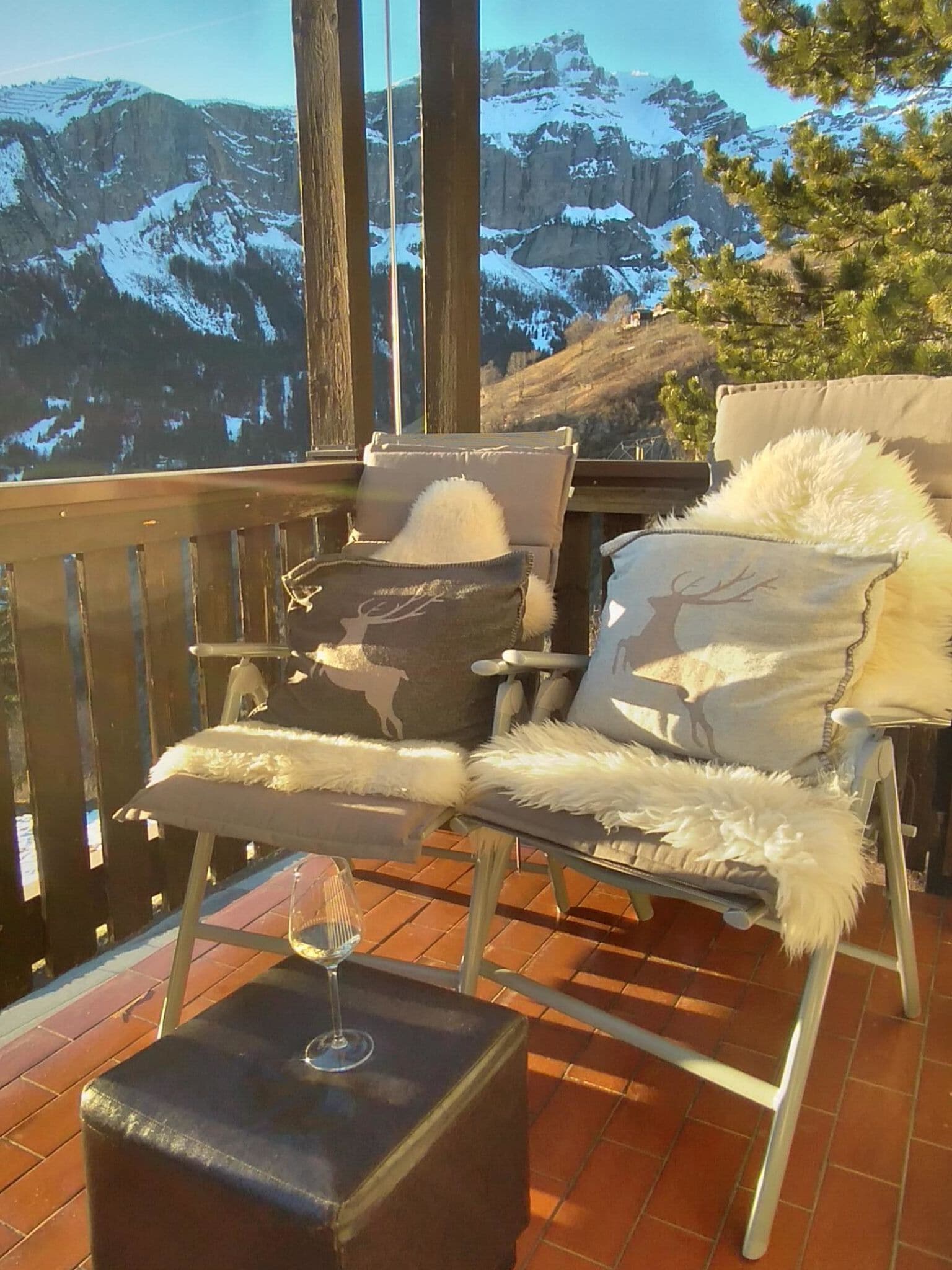 Ein Balkon mit zwei Stühlen, einer mit Kopfkissen mit Reh-Motiv und der andere mit Reh-Silhouette, sowie ein Glas Wein auf einem kleinen Tisch.