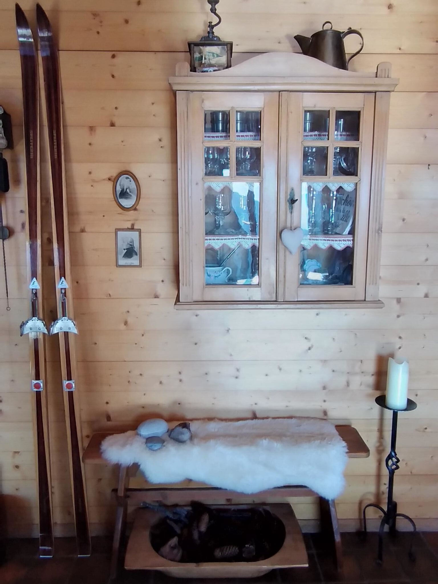 A wooden cabin interior with a wooden cabinet with glass doors, skis leaning against the wall, and a fur-covered bench.