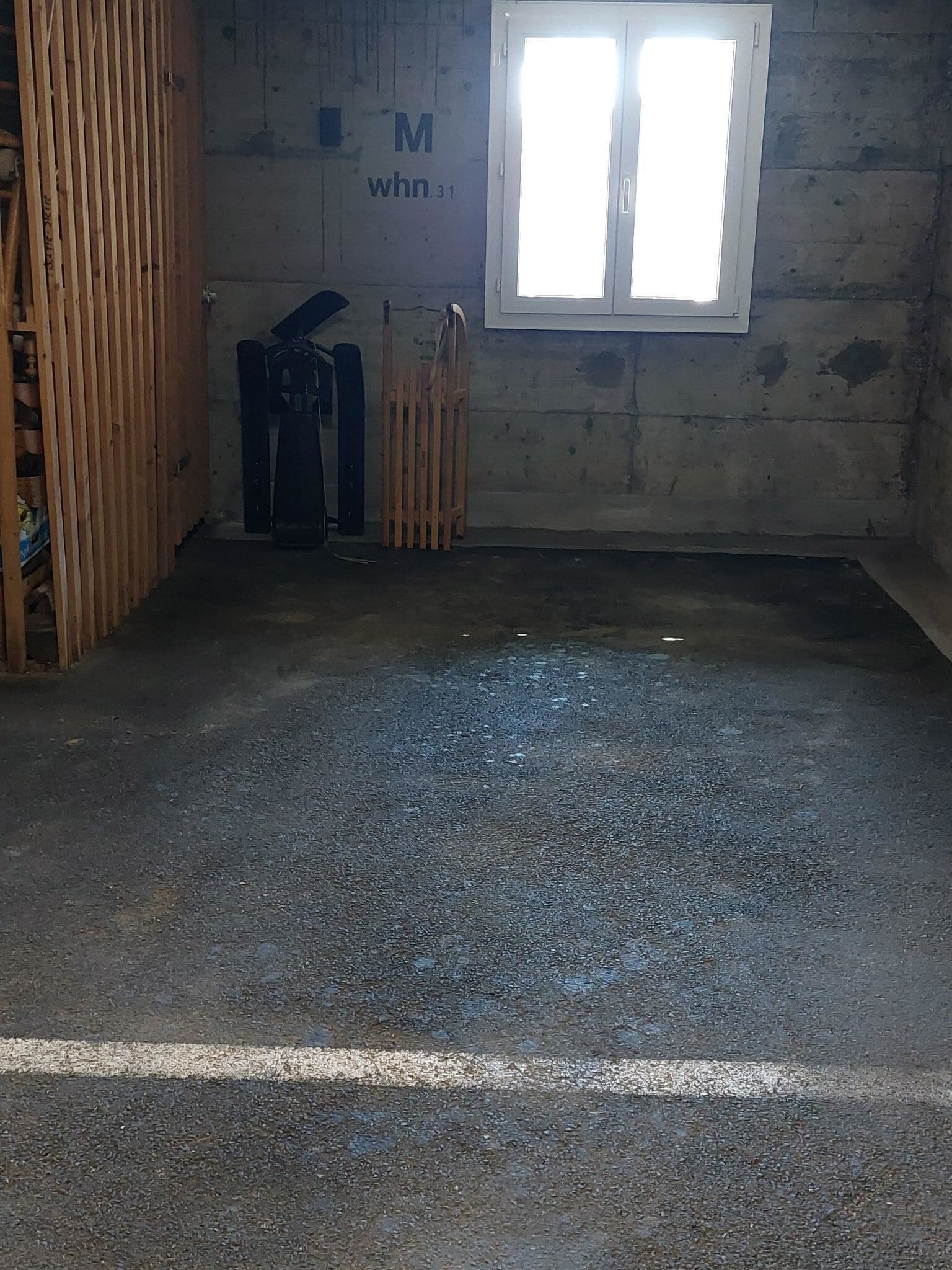Une pièce avec un sol en béton, une fenêtre aux cadres blancs et une cloison en bois portant les inscriptions « M » et « whn.31 ».