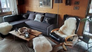 Ein gemütliches Wohnzimmer mit einem grauen Sofa, einem Holztisch, einem flauschigen weißen Pouf, einem schwarzen Ledersessel und einem rustikalen Kamin.
