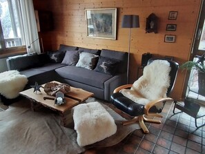 Ein gemütliches Wohnzimmer mit einem grauen Sofa, einem Holztisch, einem flauschigen weißen Pouf, einem schwarzen Ledersessel und einem rustikalen Kamin.