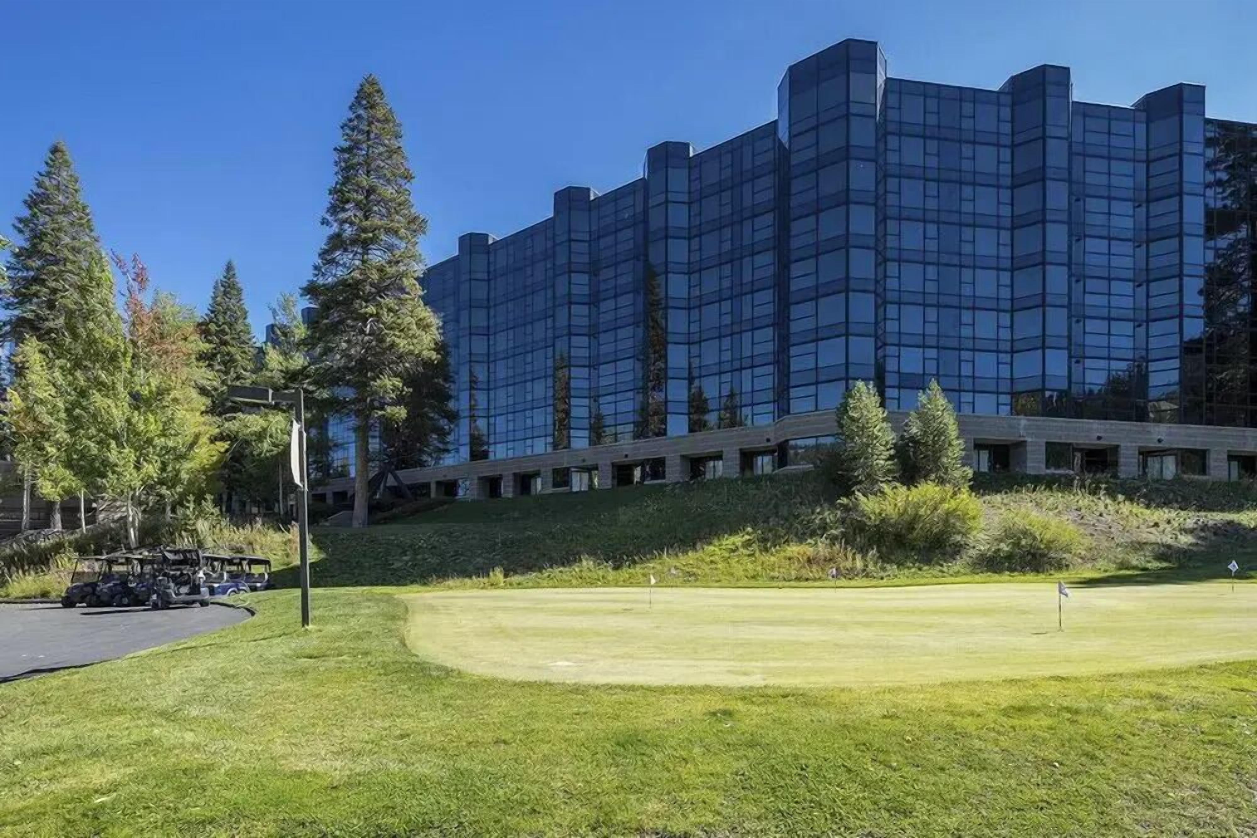 A modern building with a reflective glass facade, a well-maintained grassy area with a flag, and a parking lot with vehicles.