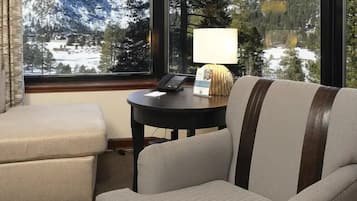 A room with a sofa, a small table with a lamp, and a view of snow-covered mountains through large windows.