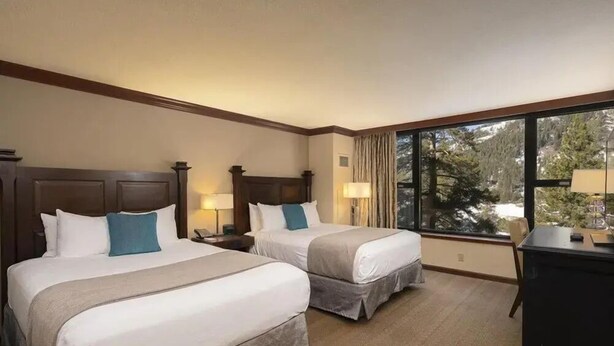 Habitación de hotel con dos camas, un escritorio y un ventanal con vistas a árboles.