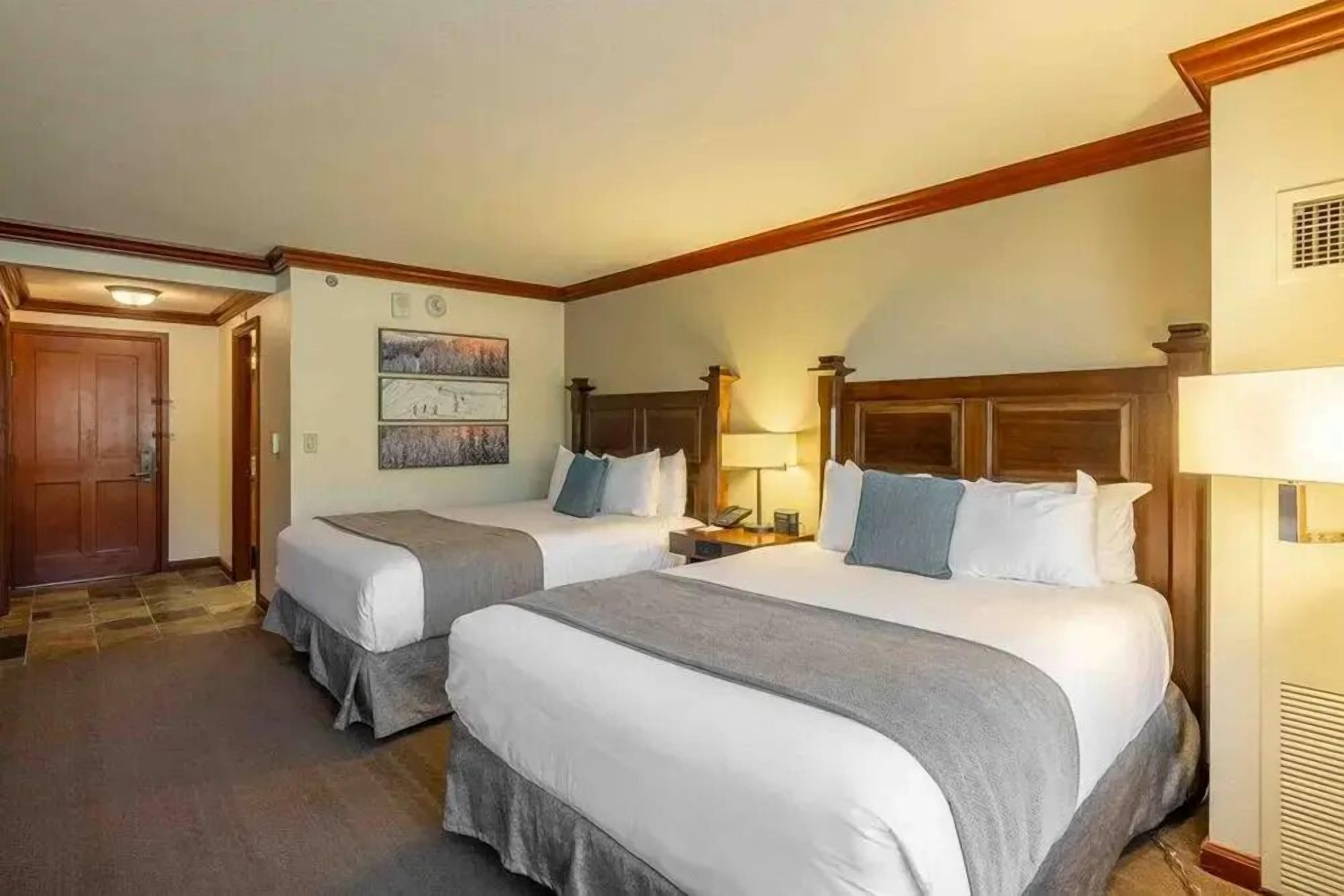A hotel room with two beds, a wooden headboard, a nightstand, a lamp, and a door.