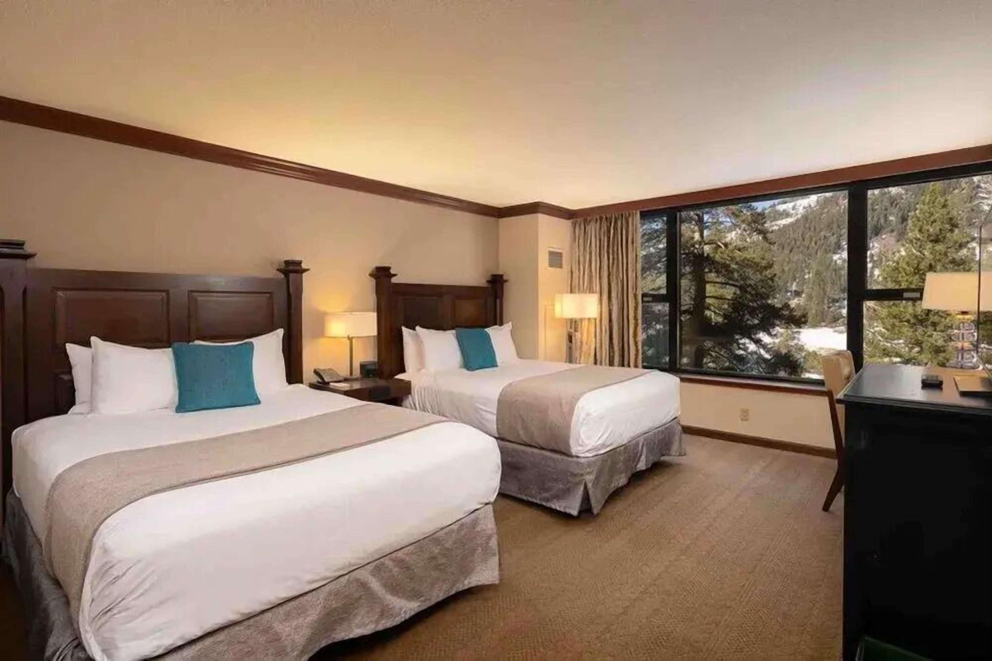 A hotel room with two beds, a desk, and a large window offering a view of trees.