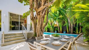 Uma área de refeições ao ar livre com piscina, cercada por plantas tropicais e uma grande árvore.
