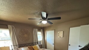A bedroom with a bed, a ceiling fan, a window with curtains, a wall decoration, a door, and a television.