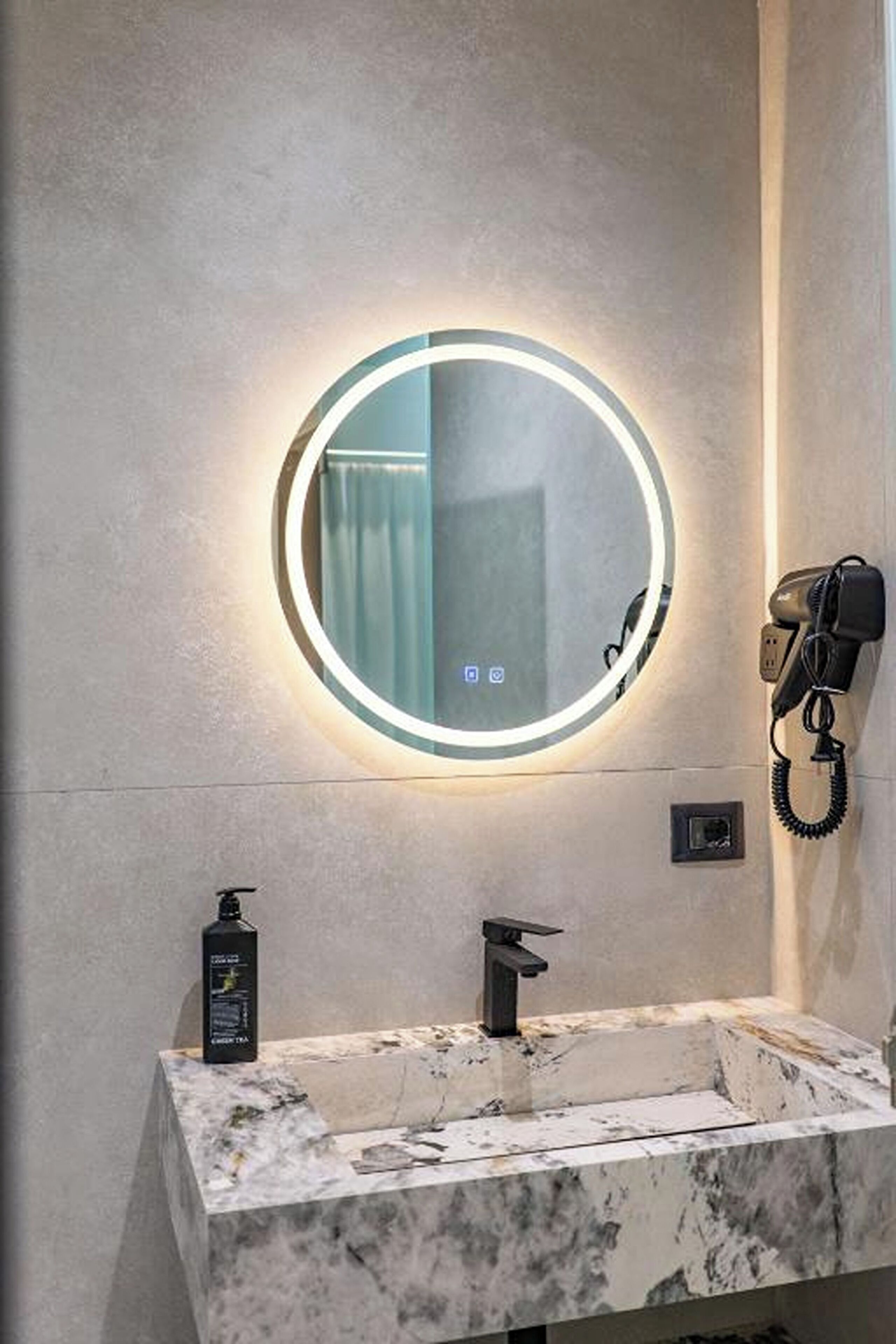 Uma casa de banho com lavatório de mármore, um espelho redondo com luz e um secador de cabelo fixado na parede.