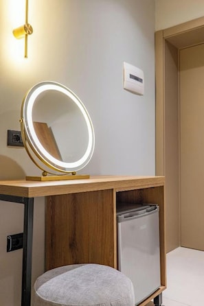 Un baño con tocador, espejo redondo con luces, taburete y cubo de basura.
