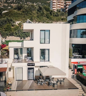 Un edificio moderno con techo plano, grandes ventanales y un área de estar exterior cubierta con mesas y sillas.