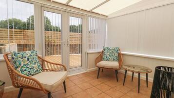 A sunlit patio with wicker chairs and a small round table, large sliding glass doors leading to an outdoor area, and a tiled floor.