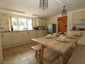 A kitchen with a wooden table set for dining, white cabinets, and a window with curtains. - Greenwich Cottage (Bridport)