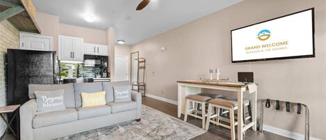 A modern living room with a sofa, a coffee table, a dining area with stools, a ceiling fan, and a kitchen in the background.