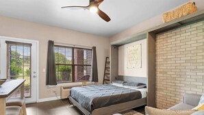 Un dormitorio con cama, escritorio, silla, ventilador de techo y pared de ladrillo.