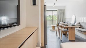 Ett modernt hotellrum med en platt-TV, ett skrivbord i trä, ett matbord med stolar, en soffa och ett stort fönster.