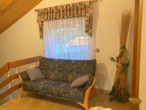 A room with a wooden-framed sofa, a window with patterned curtains, and a decorative plant. - Chalet Kristall # 6-bed apartment, Dusche, WC 6-bed apartment, Dusche, WC by Interhome (Albinen)