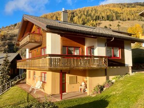 A two-story chalet with a wooden balcony, a patio with outdoor seating, and a view of a forested hillside. - Chalet Kristall # 6-bed apartment, Dusche, WC 6-bed apartment, Dusche, WC by Interhome (Albinen)