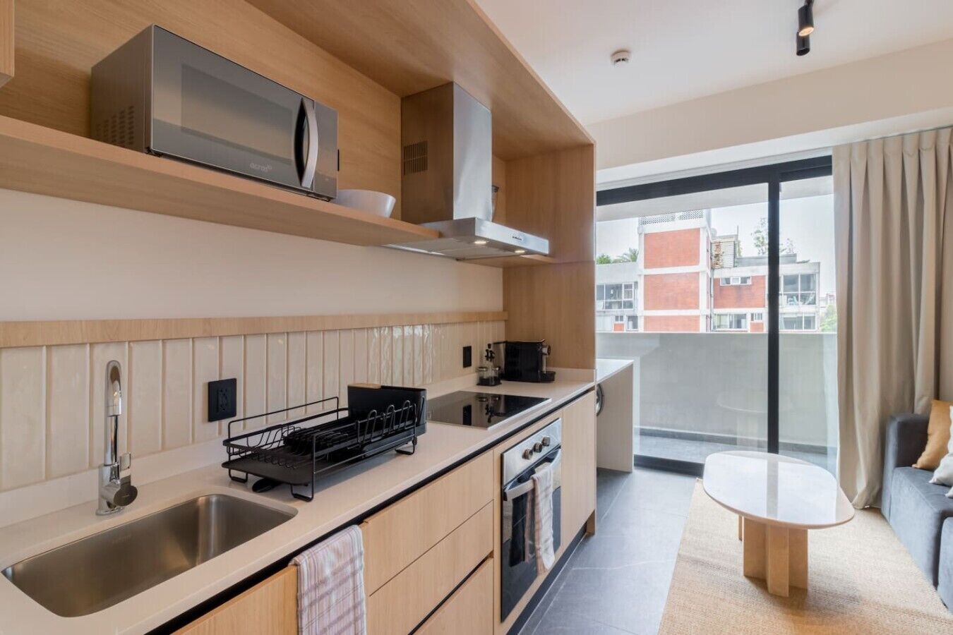 Una cocina moderna con microondas, pileta y estufa, un pequeño comedor con mesa, y vista a edificios a través de un ventanal grande.