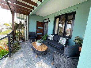 Ein Balkon mit einer Holz-Pergola, einem schwarzen Sofa mit floralen Kissen, einem runden Holztisch und Topfpflanzen.