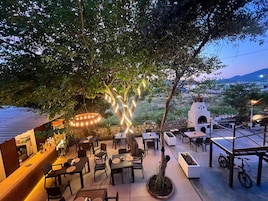 An outdoor dining area with tables and chairs, a stone oven, and a tree providing shade.