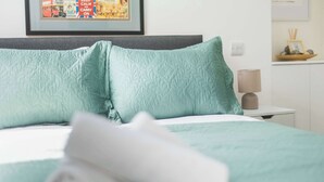 Een net opgemaakt bed met een opgerolde witte handdoek, een kussentje in teal-kleur en een ingelijst foto op de muur.