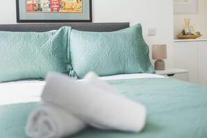 A neatly made bed with a rolled white towel, a teal quilted pillow, and a framed picture on the wall. - Spacious 2BDR 2BA near Shepherds Bush Oasis (Greater London)