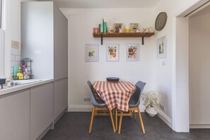 A compact kitchen with a dining table, chairs, a shelf with books and decorative items, and a clock on the wall. - Spacious 2BDR 2BA near Shepherds Bush Oasis (Greater London)