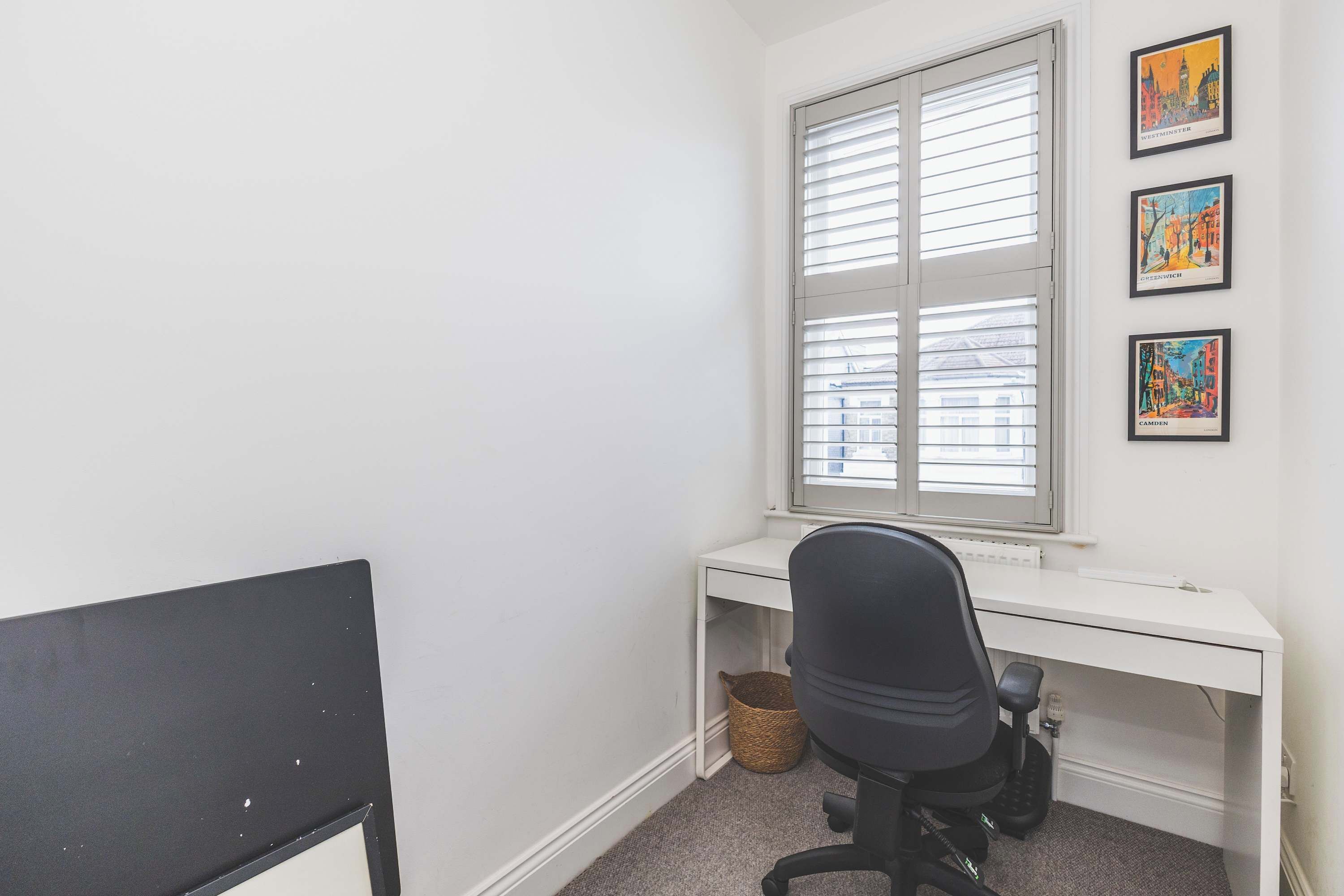 A room with a white desk, a black office chair, a window with blinds, and three framed pictures on the wall.