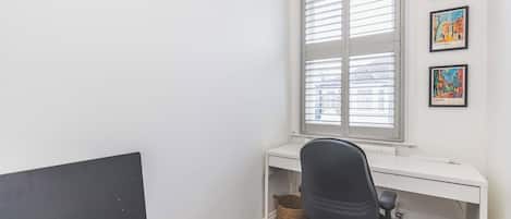 Une pièce avec un bureau blanc, un fauteuil de bureau noir, une fenêtre avec des stores et trois tableaux encadrés accrochés au mur.