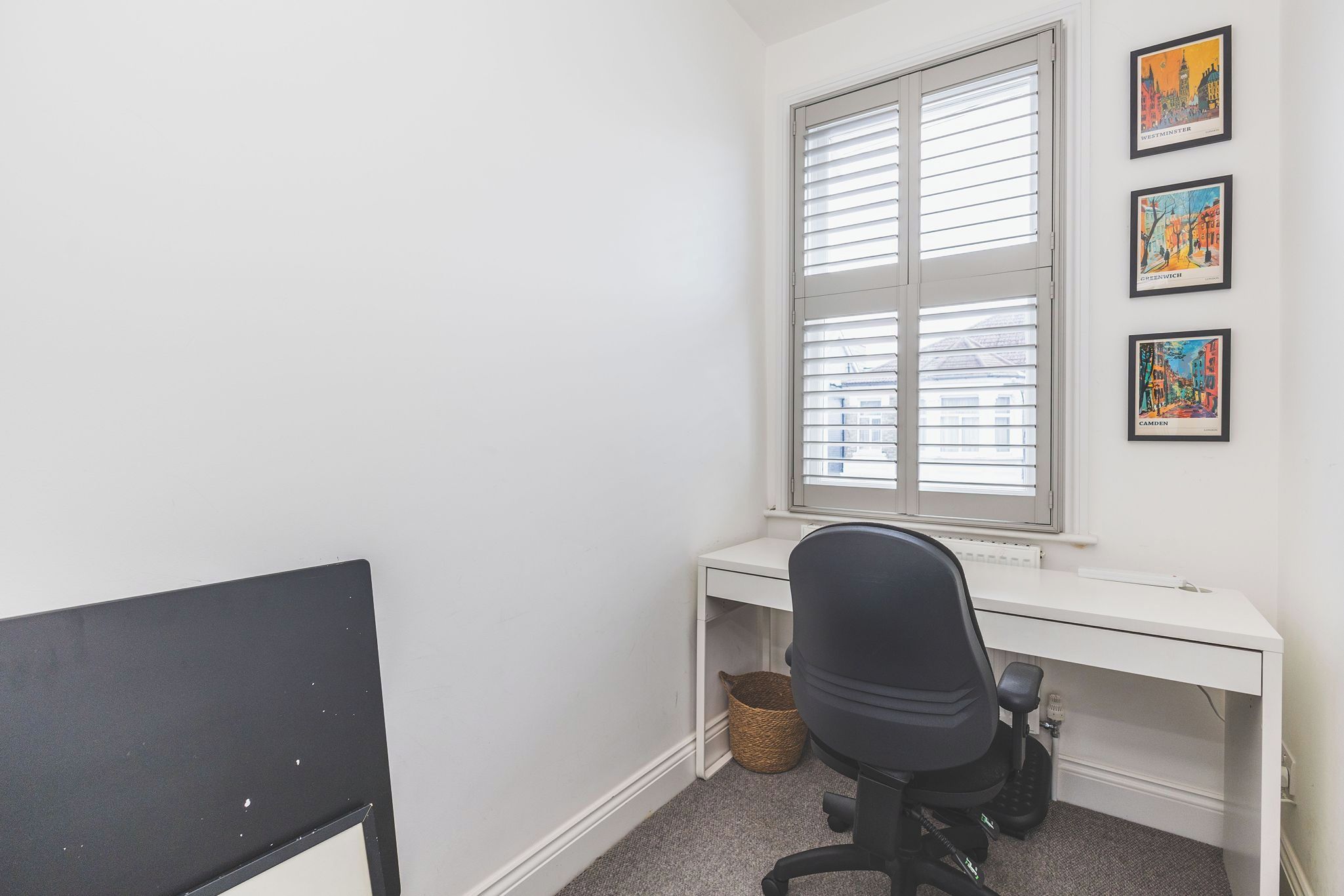 Una habitación con un escritorio blanco, una silla de oficina negra, una ventana con persianas y tres cuadros en la pared.