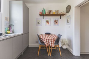 A compact kitchen with a dining area, featuring a checkered tablecloth, a bowl, and a shelf with books and decorative items. - Spacious 2BDR 2BA near Shepherds Bush Oasis (Greater London)