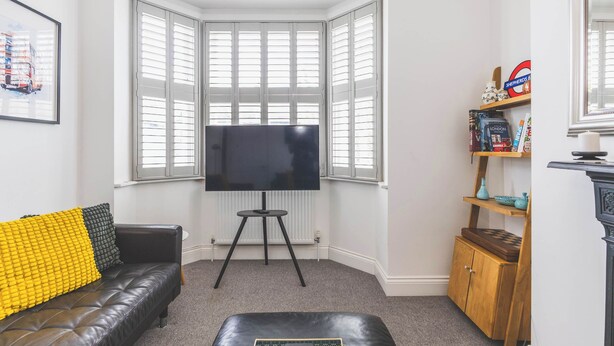 Een woonkamer met een zwarte leren bank, een zwart voetenbankje, een flatscreen televisie op een standaard, een boekenplank met boeken en decoratieve objecten, en een hoekraam met witte luiken.