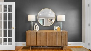 A hallway with a dark wall, a wooden console table with two lamps, a round mirror, and a framed welcome sign. - Main House + Studio Cottage • 2 Acres (Greer)