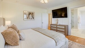A bedroom with a bed, a dresser, a television mounted on the wall, and a door leading to a bathroom. - Main House + Studio Cottage • 2 Acres (Greer)