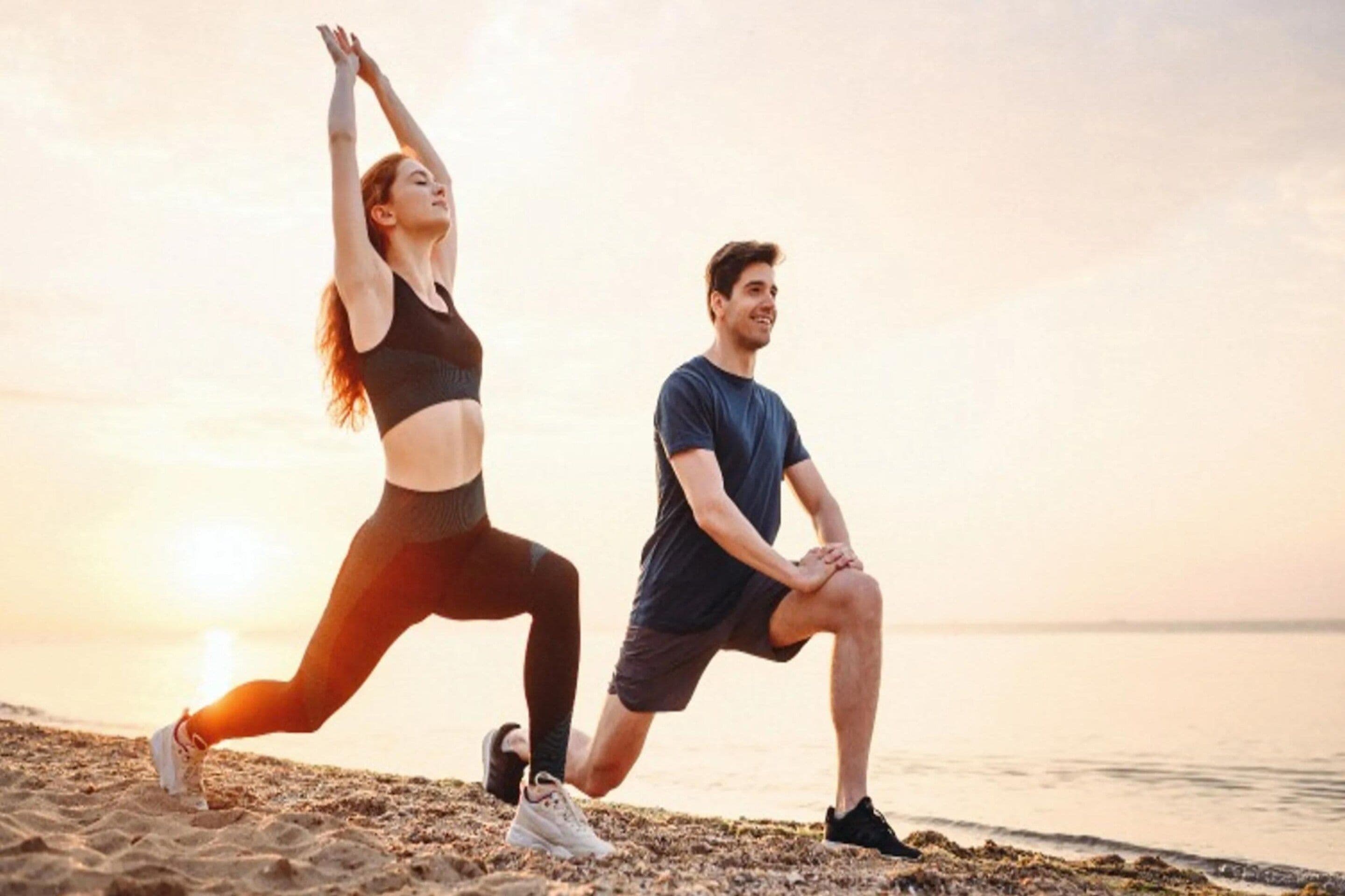 Two people performing stretching exercises on a beach with the ocean in the background.