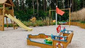Une structure de jeux en bois, dotée d’un toboggan et de balançoires, entourée de sable et de verdure.