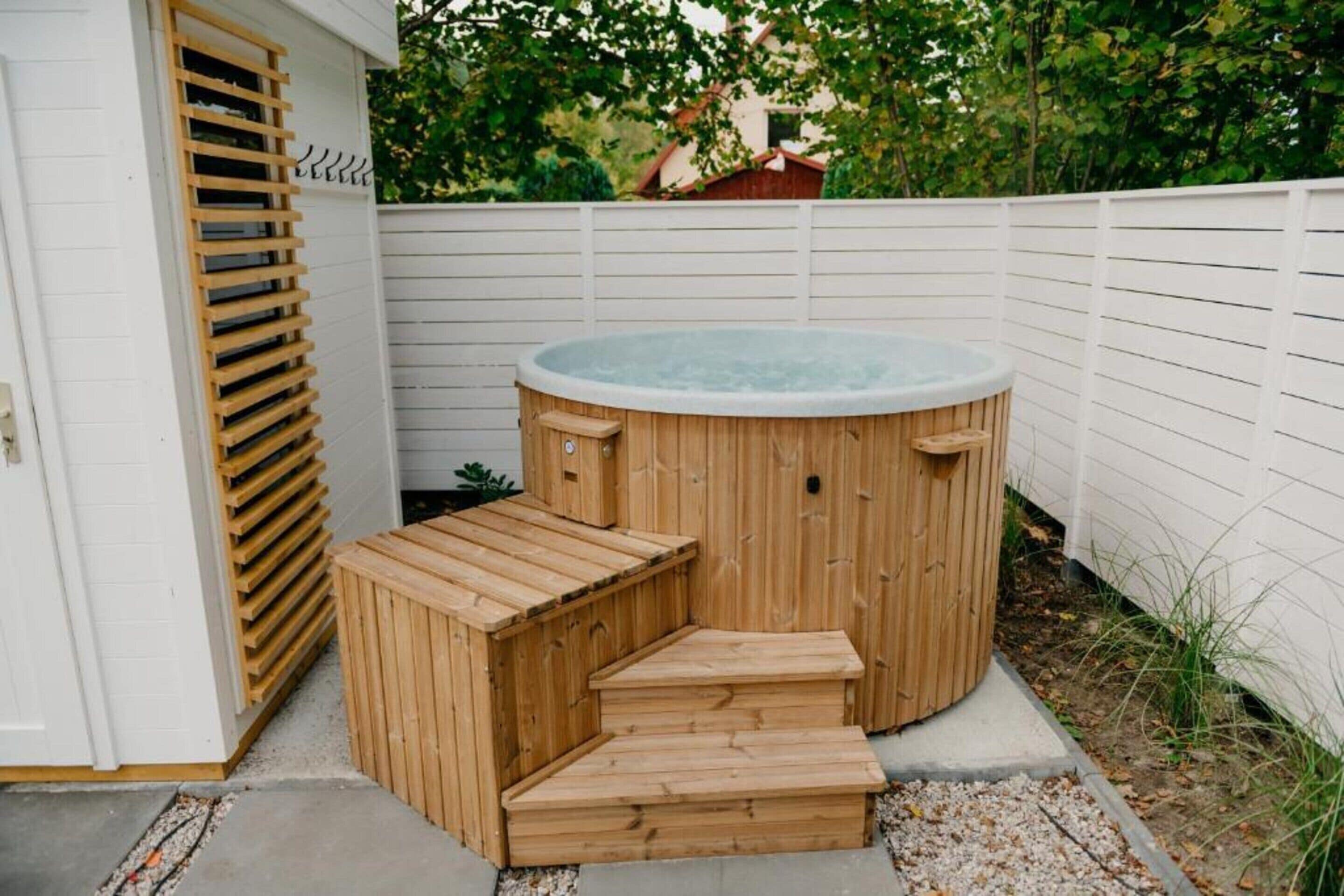 A wooden hot tub with steps, a white fence, and greenery in the background.