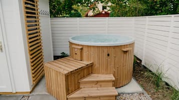 A wooden hot tub with steps, a white fence, and greenery in the background.