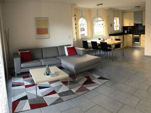 A modern living room with a grey sofa, a dining table, and a kitchen area with cabinets and appliances.