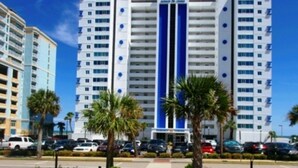 A multi-story hotel with a central blue stripe, surrounded by palm trees and parked cars. - Cozy 2-bedroom Oceanfront Family Friendly Condo (Myrtle Beach)