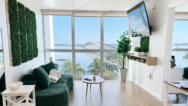 A modern living room with a green sofa, a small round table, a TV mounted on the wall, and a view of a bridge and water. - Waterfront Retreat in Downtown Fort Myers (Fort Myers)