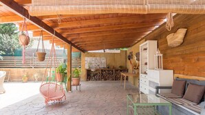 Een overdekt terras met stenen vloer, een hangstoel, een bank, een tafel en een keuken met fornuis en kastjes.