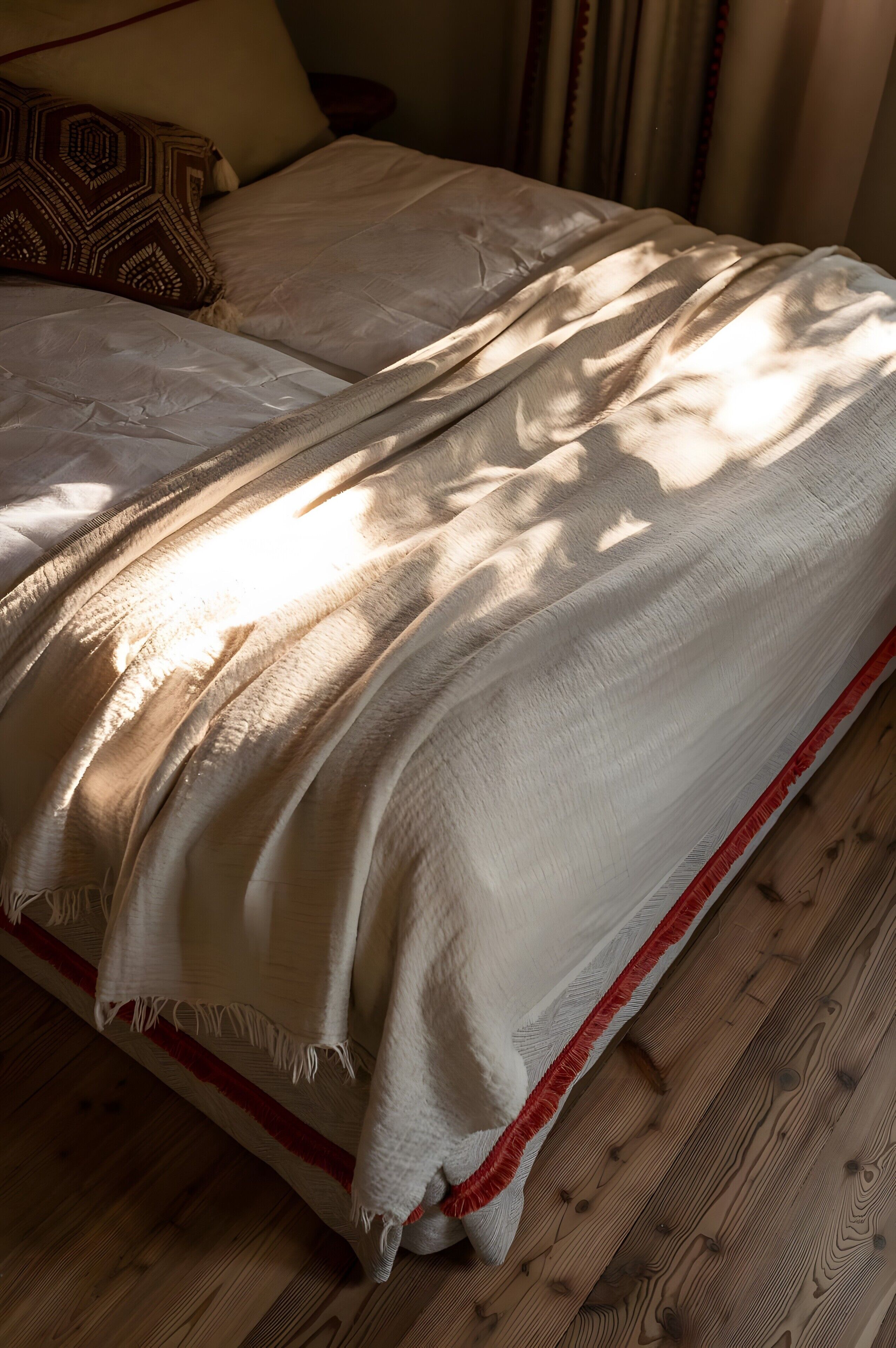 A bed with a white blanket and a red trim, wooden flooring, and a patterned pillow.
