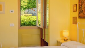 A room with a bed, a bedside table with a lamp, a window with a view of greenery, and a door. - Corte Pulchra - La Lavanda 2 (Pozzolengo)