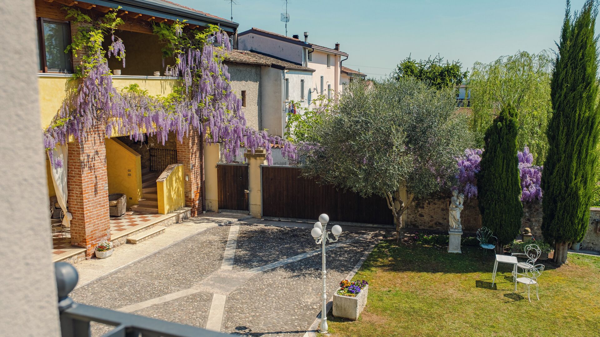 Une cour avec un escalier, une pergola ornée de fleurs violettes, une statue et un espace aménagé pour s’asseoir.